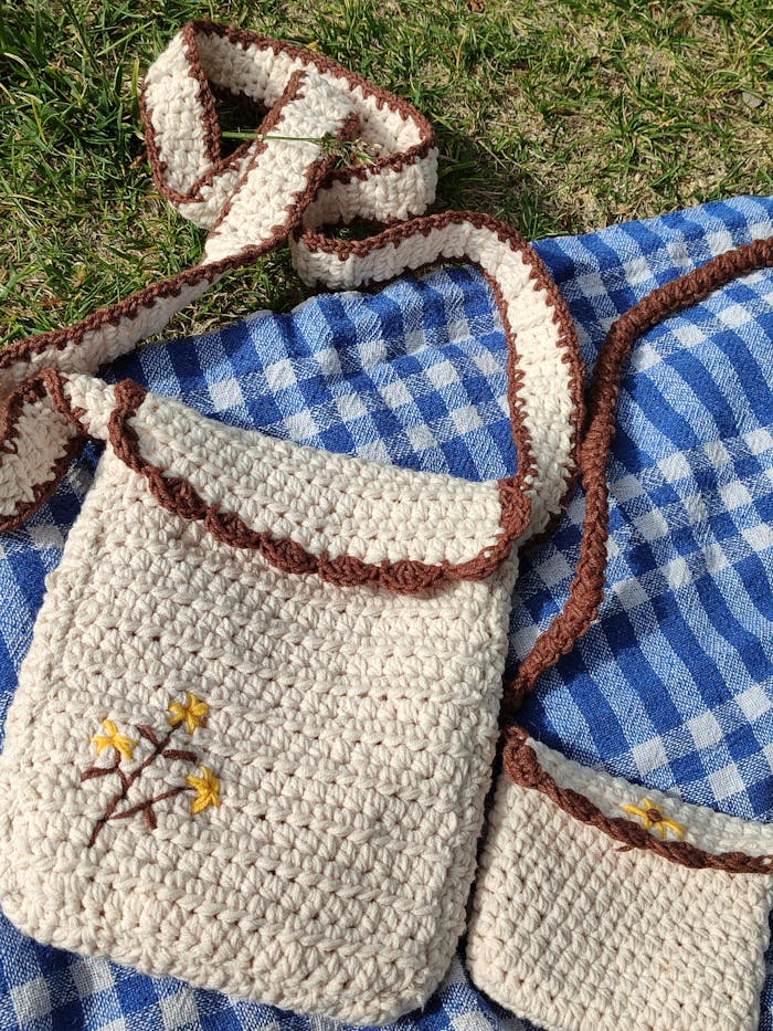 A crochet bag with floral embroidery rests on a checkered picnic blanket outdoors.